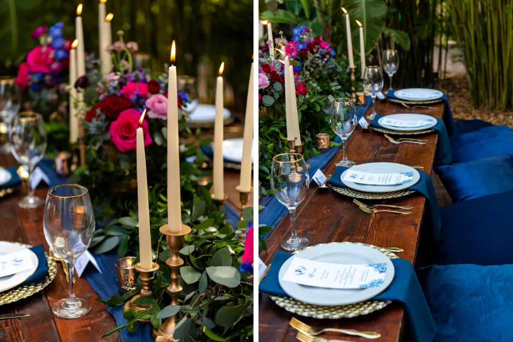 Wedding Reception Decor, Long Wooden Table with Velvet Blue Pillows, Blue Table Runner and Linens, Pale Blue Plates, Gold Silverware, Small and Tall Gold Candlesticks, Greenery Garland, Pink, Blue, and Red Low Floral Centerpieces, Bamboo Garden Backdrop | St. Pete Wedding Venue NOVA 535 | Tampa Bay Wedding Photographer Caroline and Evan Photography | Florist Monarch Events and Design | Designer and Planner Southern Weddings and Events | Rentals A Chair Affair
