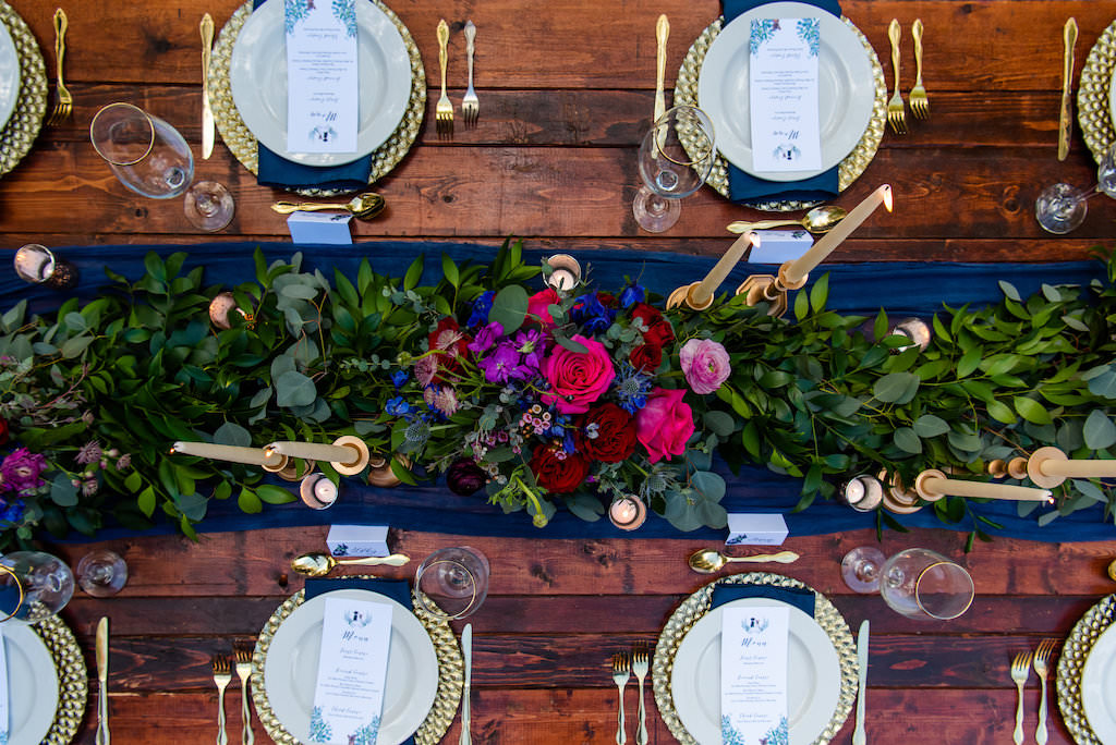 Wedding Reception Decor, Long Wooden Table with Velvet Blue Pillows, Blue Table Runner and Linens, Pale Blue Plates, Gold Silverware, Small and Tall Gold Candlesticks, Greenery Garland, Pink, Blue, and Red Low Floral Centerpieces, Bamboo Garden Backdrop | St. Pete Wedding Venue NOVA 535 | Tampa Bay Wedding Photographer Caroline and Evan Photography | Florist Monarch Events and Design | Designer and Planner Southern Weddings and Events | Rentals A Chair Affair