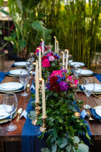 Wedding Reception Decor, Long Wooden Table with Velvet Blue Pillows, Blue Table Runner and Linens, Pale Blue Plates, Gold Silverware, Small and Tall Gold Candlesticks, Greenery Garland, Pink, Blue, and Red Low Floral Centerpieces, Bamboo Garden Backdrop | St. Pete Wedding Venue NOVA 535 | Tampa Bay Wedding Photographer Caroline and Evan Photography | Florist Monarch Events and Design | Designer and Planner Southern Weddings and Events | Rentals A Chair Affair