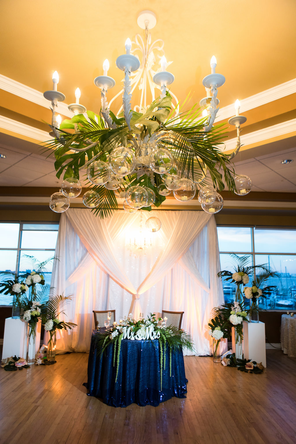 Classic Traditional Tropical Inspired Ballroom Wedding Reception Decor, White Chandelier with Hanging Clear Glass Balls, Palm Tree Leaves, Sweetheart Table with Sparkle Blue Tablecloth, Green Hanging Amaranthus, White Mr and Mrs Sign, White Drapery Backdrop, White Pedestals with Tropical Floral Bouquets | St. Petersburg Wedding Venue Isla Del Sol Yacht and Country Club