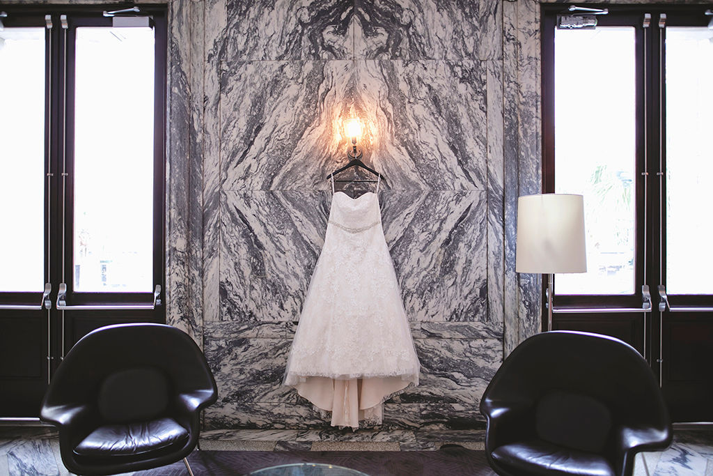 Strapless Lace Ballgown Wedding Dress with Rhinestone Belt Hanging in Front of Black and White Marble Wall