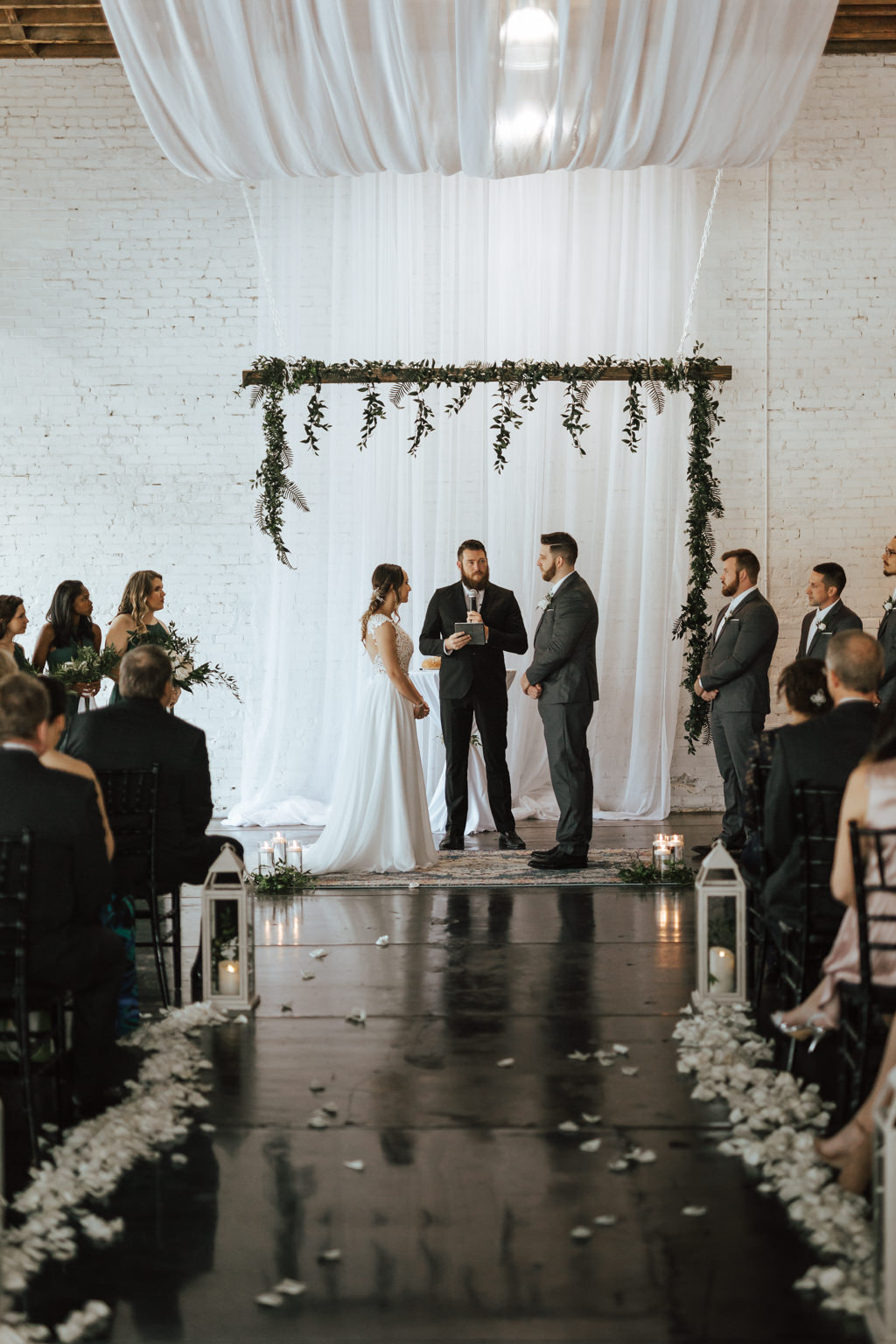 Romantic Wedding Ceremony Decor, Bride and Groom Exchanging Vows Under Greenery Arch and White Draping Wedding Portrait | Lakeland Industrial Wedding Venue HAUS 820