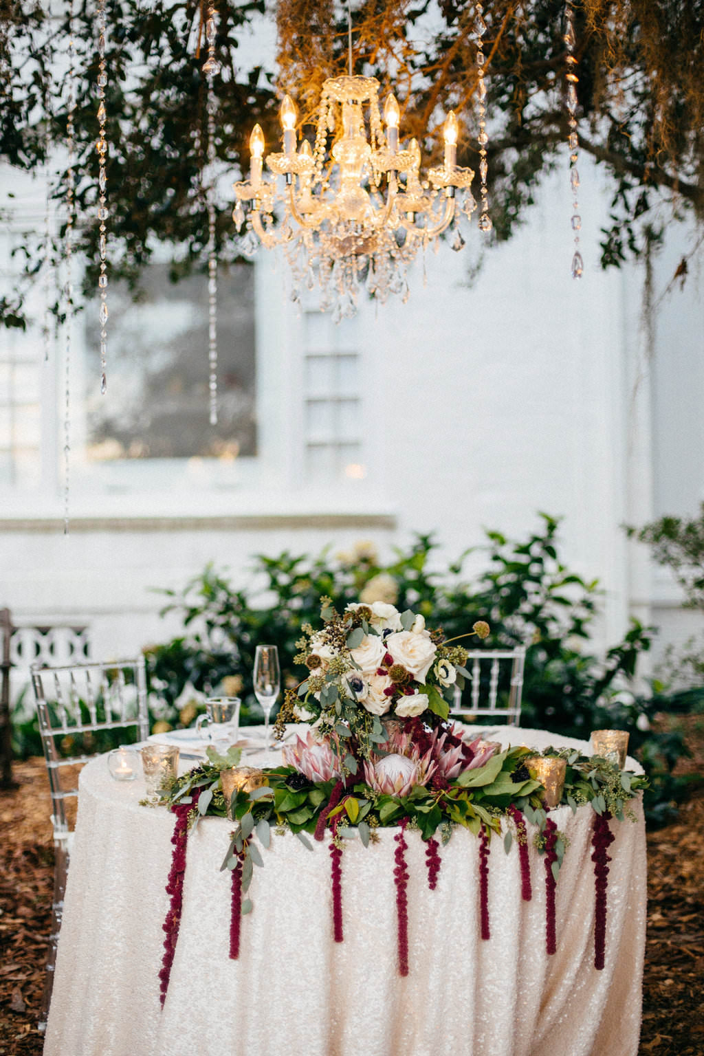 Outdoor Garden Wedding Reception Decor, Sweetheart Table with Ivory Tablecloth, Greenery Garland and Red Hanging Amaranthus, Ivory Roses, White Anemones, King Protea and Greenery Flower Centerpiece, Hanging Crystal Chandelier | Tampa Bay Wedding Planner NK Productions