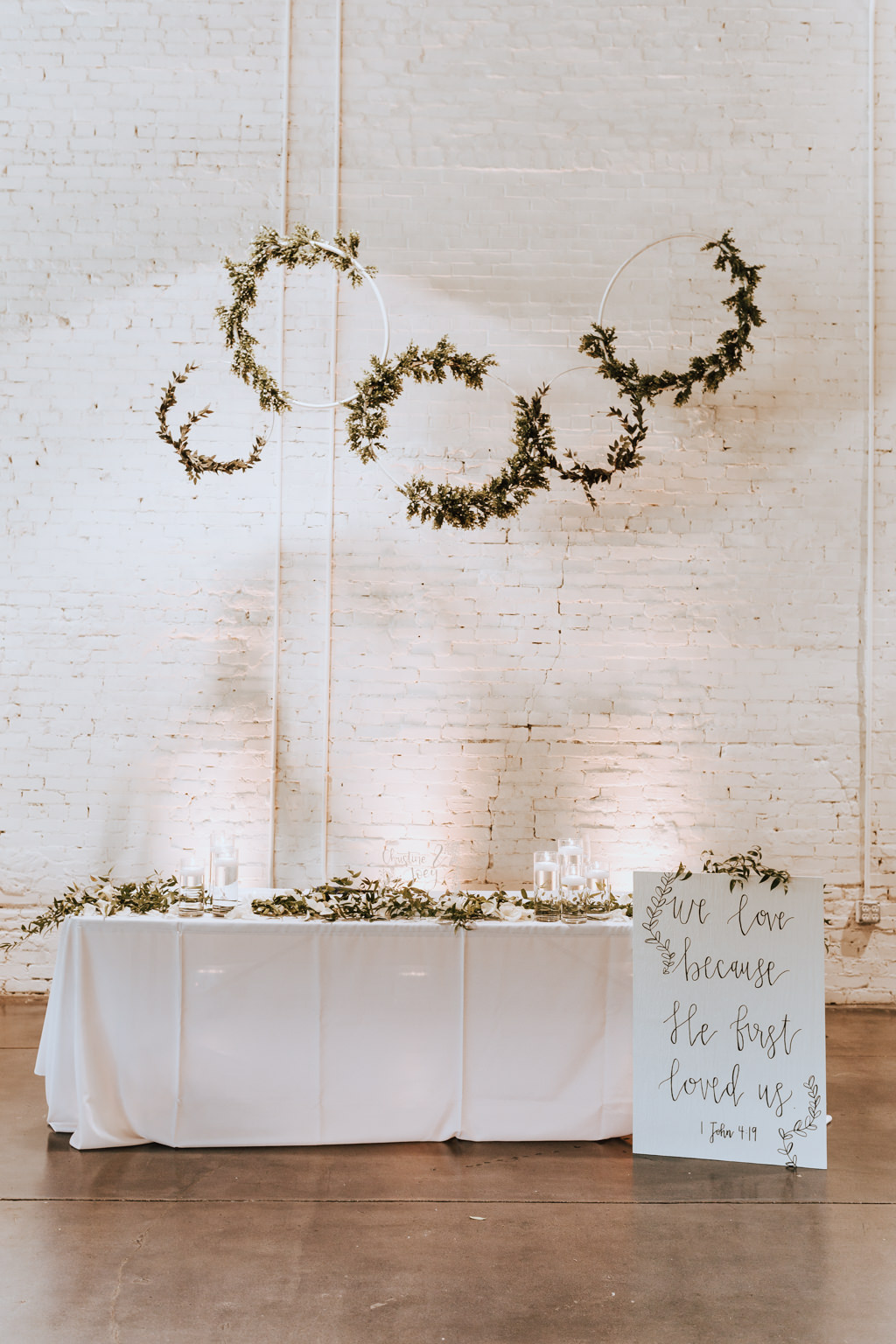 Wedding Reception Decor, Sweetheart Table with White Tablecloth, Greenery Garland, Glass Cylinders with Floating Candles, White Wedding Sign, White Brick Wall Backdrop, Hanging Rings with Greenery Garland | Lakeland Industrial Wedding Venue HAUS 820