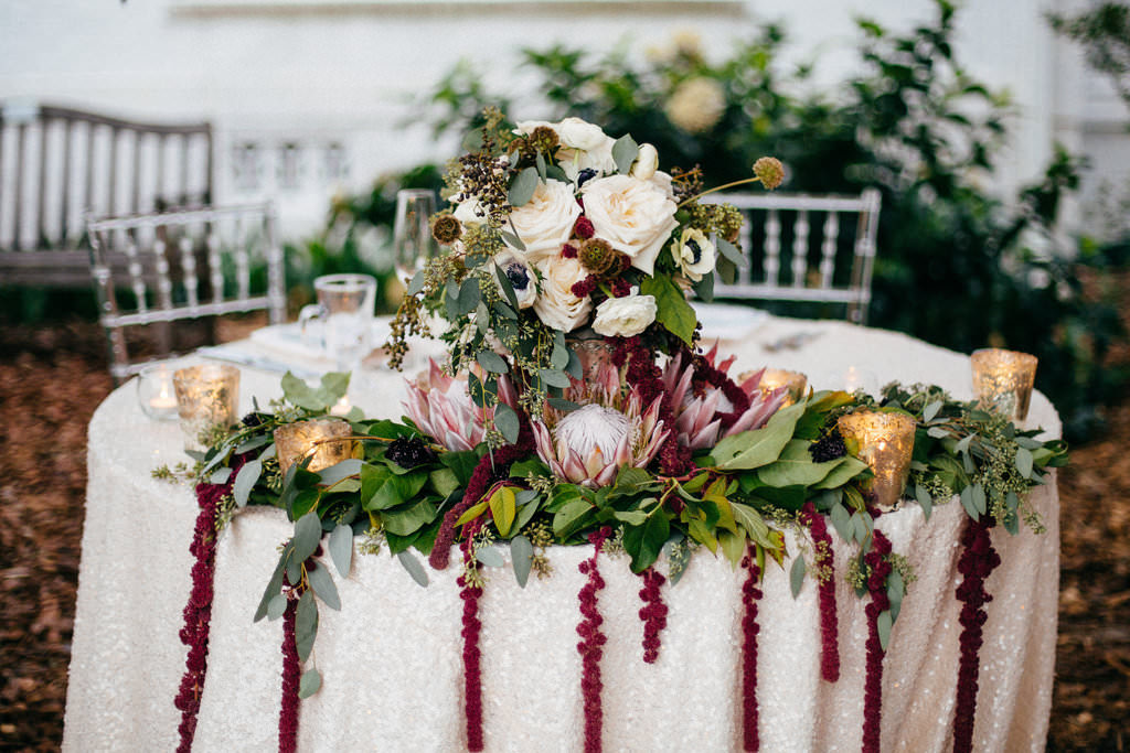 Outdoor Garden Wedding Reception Decor, Sweetheart Table with Ivory Tablecloth, Greenery Garland and Red Hanging Amaranthus, Ivory Roses, White Anemones, King Protea and Greenery Flower Centerpiece, Gold Mercury Candle Votives | Tampa Bay Wedding Planner NK Productions