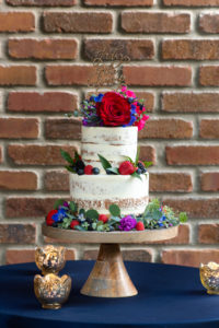 Two Tier Rustic Semi Naked Wedding Cake with Red, Purple, Pink, Blue and Greenery Real Flower Decor, Laser Cut Cake Topper and Red Rose, Pink Floral Cake Topper on Wooden Cake Stand, Blue Tablecloth, Gold Mercury Candles | Tampa Bay Wedding Photographer Caroline and Evan Photography | Florist Monarch Events and Design | Designer and Planner Southern Glam Weddings and Events | Cake Baker Artistic Whisk