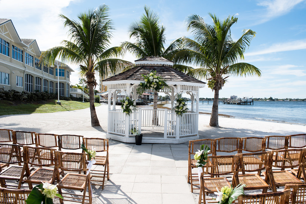 Outdoor Beach Wedding Ceremony at Gazebo with White Florals and Green Palm Tree Branches, Wooden Folding Chairs | Waterfront Venue Isla Del Sol Yacht and Country Club