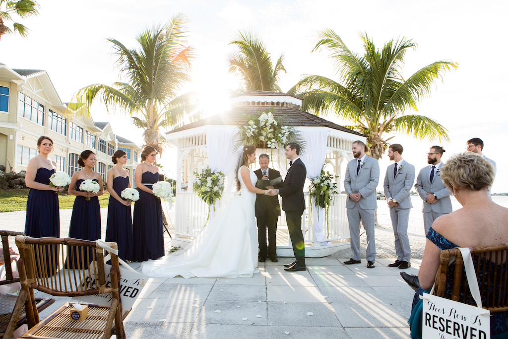 Florida Outdoor Bride and Groom Exchanging Vows During Beachfront Wedding Ceremony at Gazebo with White Drapery and White Flowers with Palm Tree Branches Decor | Waterfront Venue Isla Del Sol Yacht and Country Club