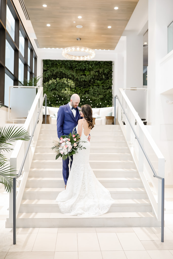 Destination Florida Bride and Groom Wedding Portrait on Hotel Staircase in Courtyard, Groom in Blue Tuxedo, Bride in White and Pink Flower Bouquet with Green Palms, Bride in Spaghetti Strap Backless White Fitted Hayley Paige Wedding Dress | Photographer Lifelong Photography Studios | Hotel Wedding Venue The Hilton Clearwater Beach Resort & Spa