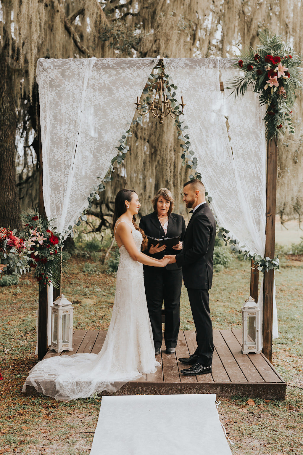 Bride and Groom Wedding Ceremony Under Romantic Wooden Wedding Ceremony Arch on Platform with Lace Drapes, Green Garland, Rich Red and Pink Floral Arrangements with Greenery, White Lanterns, on Rural Florida Farm Backdrop, Bride in Beaded White Spaghetti Strap Wedding Dress | Tampa Bay Wedding Dress Shop Truly Forever Bridal | Rustic Venue The Orange Blossom Barn