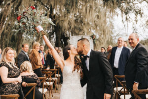 Bride and Groom Intimate Wedding Portrait, Kissing During Wedding Recessional While Holding Red and Green Floral Wedding Bouquet in Rustic Tampa Bay Wedding Venue