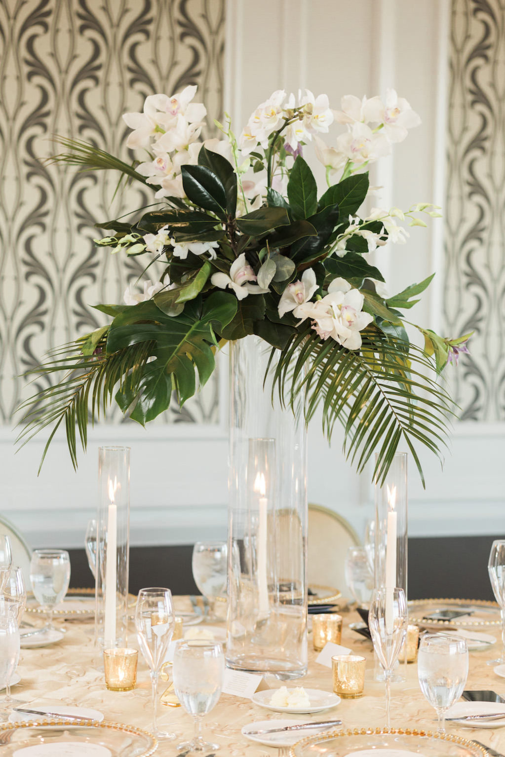 Classic, Tropical Inspired Wedding Reception Decor, Tall Glass Cylinder Vase with Palm Tree Branches, White Orchids and Green Leaves Floral Centerpiece, Round Table with Gold Tablecloth, Tall Candlesticks in Clear Cylinder Vases