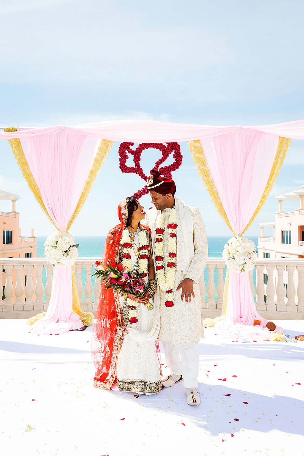 Florida Indian Bride and Groom Wedding Ceremony Portrait, Blush Pink and Gold Linen Drapery Arch, White and Blush Pink Floral Bouquets, Bride Wearing White Saree and Red Headdress, Groom Wearing White Dress Suit and Red and White Floral Leis | Clearwater Beach Waterfront Wedding Venue Hyatt Regency Clearwater Beach
