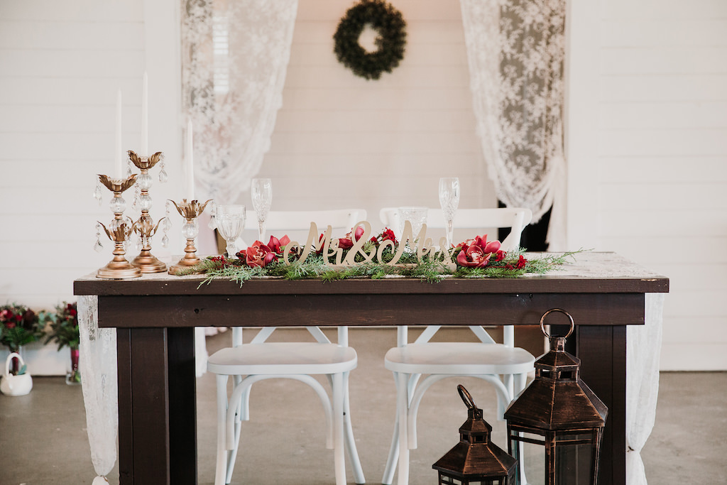 Romantic Christmas Inspired Wedding Decor at Rustic Tampa Bay Wedding, Wooden Sweetheart Table, Lace Runner, Crystal Flutes, Mr. & Mrs. Gold Table Sign, Gold Candelabra, White Candlesticks, Greenery with Red Florals, Back drop of White Draping, Green Wreath Hanging on White Shiplap Wall