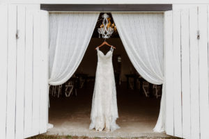 Vintage Lace Spaghetti Strap Sweetheart Neckline Lace Fitted Wedding Dress on Wooden Hanger, Against Barn Door, Lace Draping and White Chandelier Backdrop | Tampa Bay Wedding Dress Shop Truly Forever Bridal