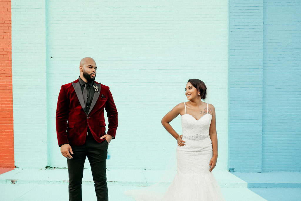 Florida Bride and Groom Wedding Portrait with Colorful Brick Wall Backdrop, Bride in Spaghetti Strap Sweetheart Neckline Lace Mermaid and Tulle Skirt Wedding Dress with Rhinestone Belt, Groom in Red Velvet Tuxedo Jacket, Black Pants and Black Dress Shirt | Lakeland Industrial Modern Wedding Venue and Event Space Haus 820