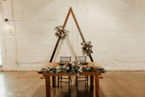 Dark and Moody Inspired Wedding Reception Decor, Triangle Arch with Dark Floral Bouquets, Wooden Sweetheart Table with Greenery Garland and Ivory Florals, Black Chiavari Chairs | Lakeland Industrial Modern Wedding Venue and Event Space Haus 820