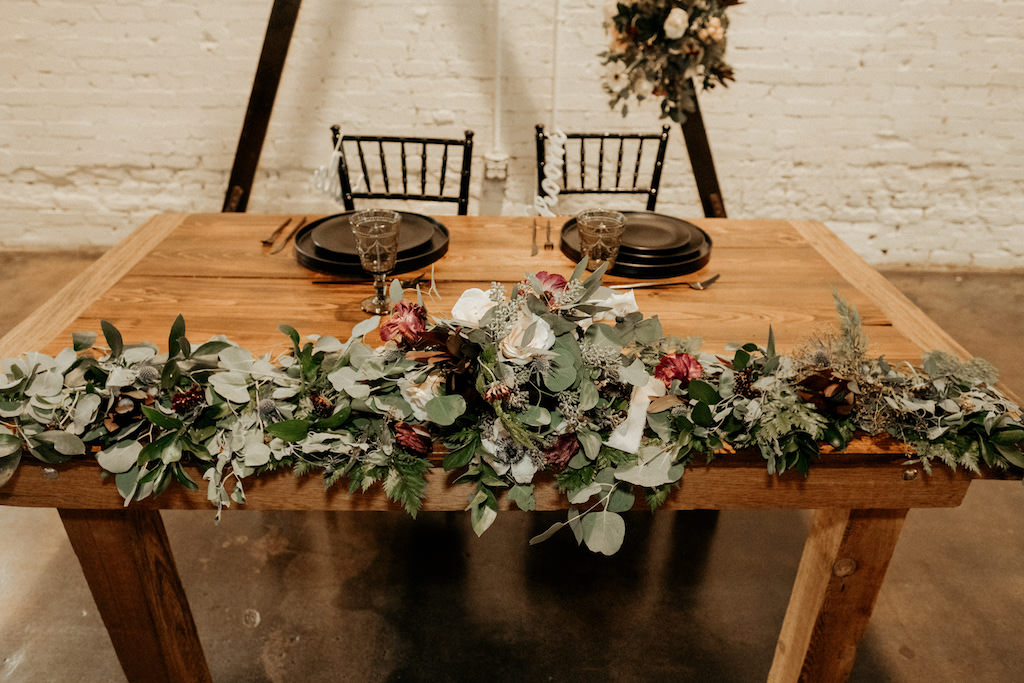 Dark and Moody Inspired Wedding Reception Decor, Wooden Sweetheart Table with Greenery Garland and Ivory and Red Florals, Black Chiavari Chairs and Black Chargers and Plates