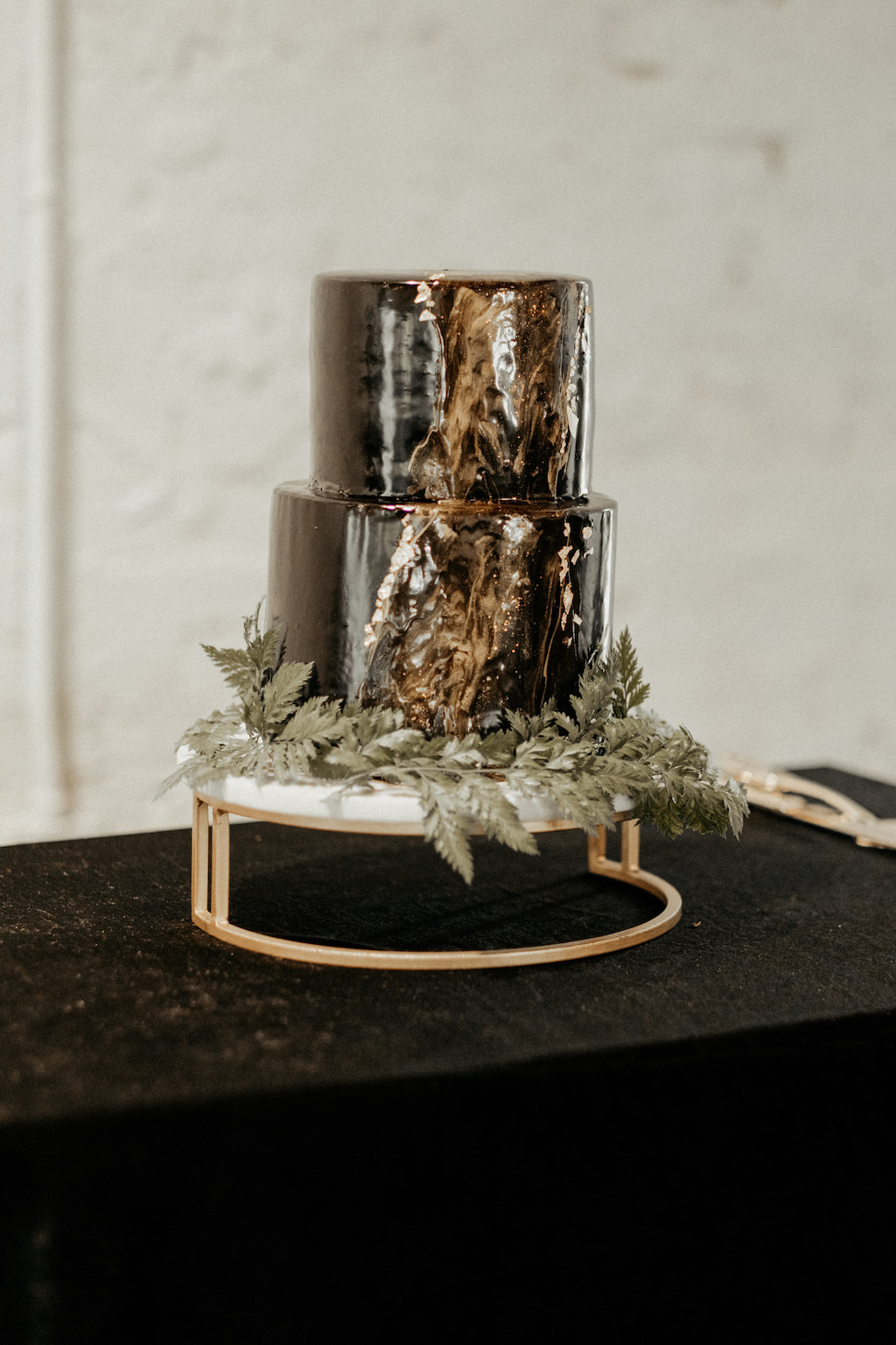Two Tier Black and Gold Marble Cake with Greenery Wreath on White and Gold Cake Stand and Black Linen Table