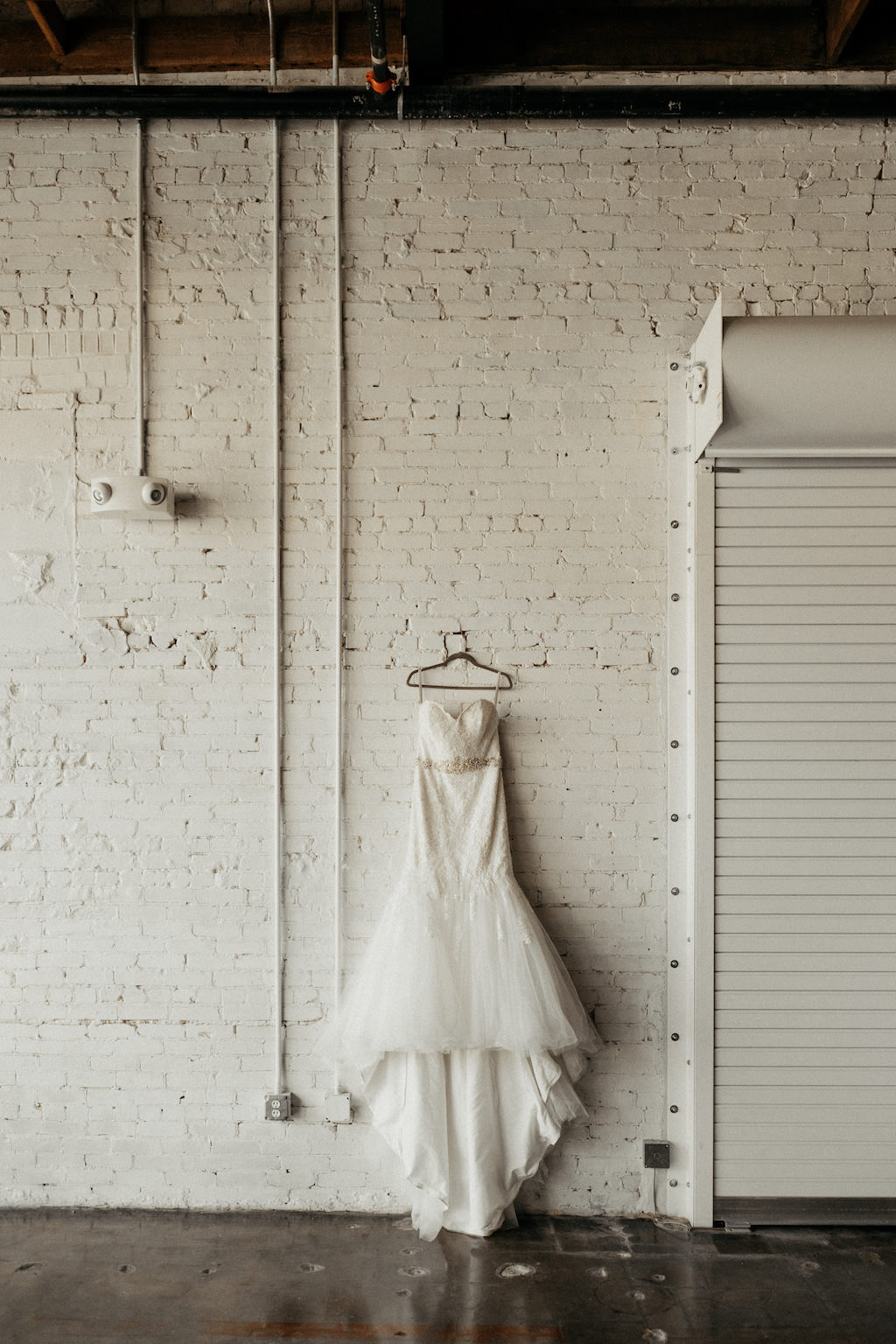 Lace Sweetheart Strapless Fitted Bodice with Rhinestone Belt, Tulle Full Skirt Wedding Dress Hanging on Hanger and White Brick Wall Backdrop | Lakeland Industrial Modern Wedding Venue and Event Space Haus 820