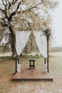 Rustic and Romantic Wooden Wedding Ceremony Arch on Platform with Lace Drapes, Green Garland, Rich Red and Pink Floral Arrangements with Greenery, White Lanterns, on Rural Tampa Bay Farm Backdrop