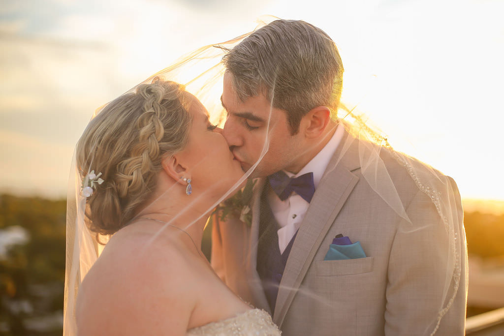 Florida Bride and Groom Kissing Underneath Veil on Rooftop Wedding Portrait at Sunset, Bridal Updo with Braid, Blue Teardrop Wedding Earrings, Groom in Light Gray Tuxedo with Blue Bowtie and Pocket Squares | Tampa Wedding Venue