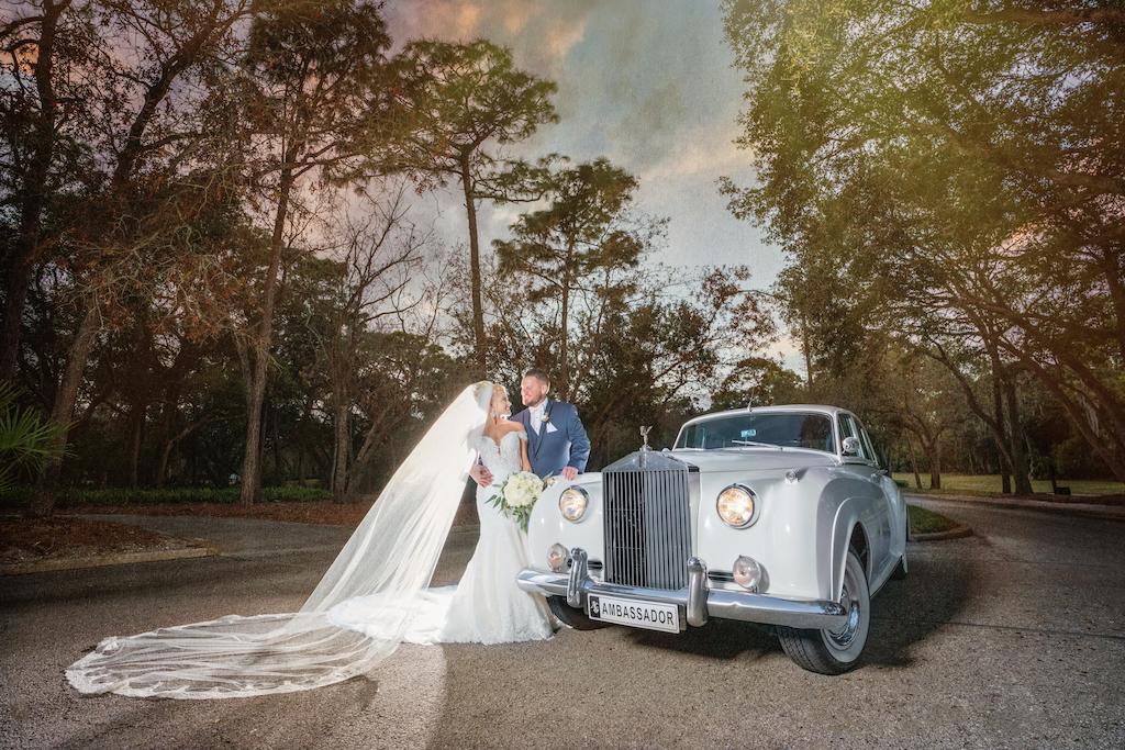 Florida Bride and Groom Wedding Portrait With Classic Bentley Car, Bride Wearing Pronovias White Lace Mermaid Style Wedding Dress With Sweetheart Neckline Fitted Bodice and Side Lace Illusion Sleeves, Veil, Carrying Elegant White Rose Bridal Bouquet with Greenery | Tampa Bay Bridal Dress Shop Nikki and Glitz Glam Boutique