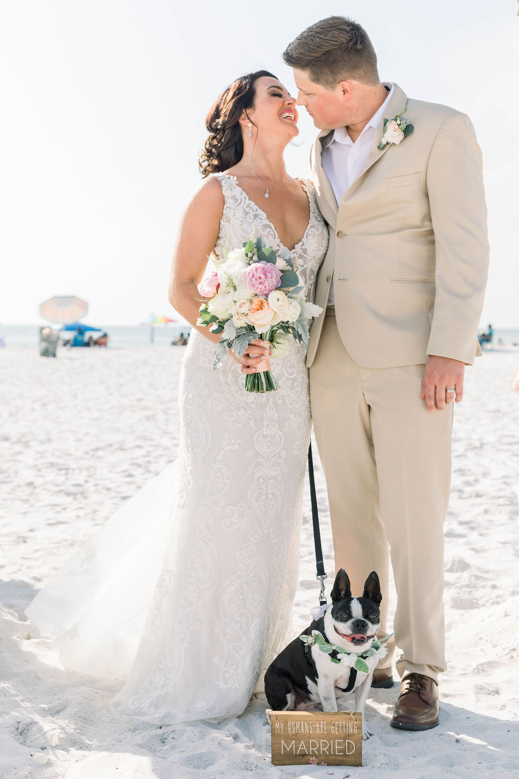 Florida Bride and Groom Wedding Portrait on Clearwater Beach with Dog, Bride in Fitted V Neckline Lace and Rhinestone Embellished Wedding Dress with Rustic Inspired Floral Bouquet, Groom in Tan Suit | Pet Planner FairyTail Pet Care | Planner Gulf Beach Weddings