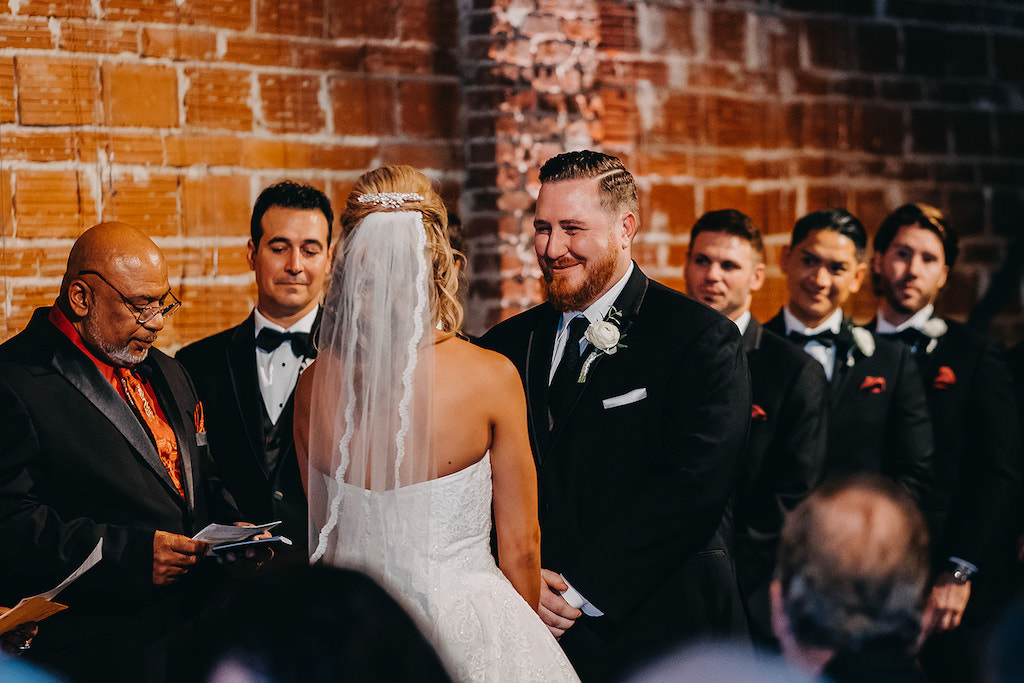 Bride and Groom at the Alter During Wedding Ceremony in Front of Red Brick Exposed Wall, Bride Wearing Veil, Groom Wearing Classic Black Tux and Tie, Groomsmen in Black Tuxedo and Black Bowtie with Red Pocket Square | Downtown St. Pete Wedding Venue NOVA 535