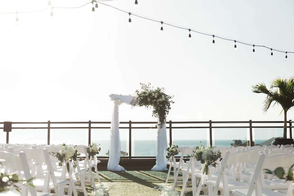 Rustic, Romantic, Beach Inspired Rooftop Waterfront Wedding Ceremony Decor , White Folding Chairs with Floral Bouquets, Arch with White Linen Drapery and Organic Ivory, Greenery Floral Arrangement | St. Pete Beach Wedding Venue Hotel Zamora