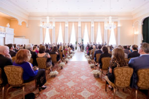Florida Bride and Groom During Ceremony in Elegant Grant Ballroom with Two Crystal Chandeliers, Long Floor to Ceiling Windows and Dropping, Armless Accent Chairs, Baby's Breath Floral Arrangement in Galvanized Metal Buckets Lining Aisle of Ceremony, South Tampa Wedding Venue Palma Ceia Golf & Country Club | Tampa Bay Wedding Planner Breezin' Weddings