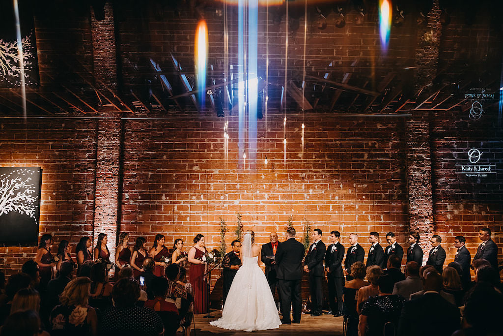 Bride and Groom with Wedding Party at the Altar During Wedding Ceremony in Front of Red Brick Exposed Wall, Ceremony Portrait with LensFlair, Bride Wearing Strapless White Ballgown with Veil, Bridesmaids in Burgundy Wine Maroon Long Dresses, Groom Wearing Classic Black Tux and Tie, Groomsmen in Black Tuxedo and Black Bowtie with Red Pocket Square | Downtown St. Pete Wedding Venue NOVA 535