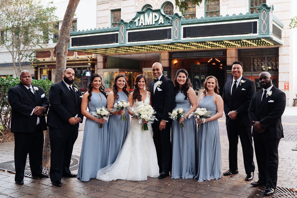 Florida Bride and Groom with Wedding Party Portrait Outside Historic Downtown Tampa Theatre, Carrying White and Ivory Floral Bouquet, Bridesmaids in Dusty Blue Halter Neckline Long Style Dresses, Groomsmen in Classic Black Tuxedo | Photographer Grind & Press Photography