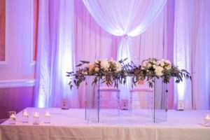 Elegant Wedding Decor at Ballroom Reception, Ghost Acrylic Sweetheart Table with Ghost Acrylic Chairs, Clear Candle Votives, Centerpiece with White Roses and Greenery, White Draping and Backlighting | Tampa Bay Wedding Venue Innisbrook Golf & Spa Resort