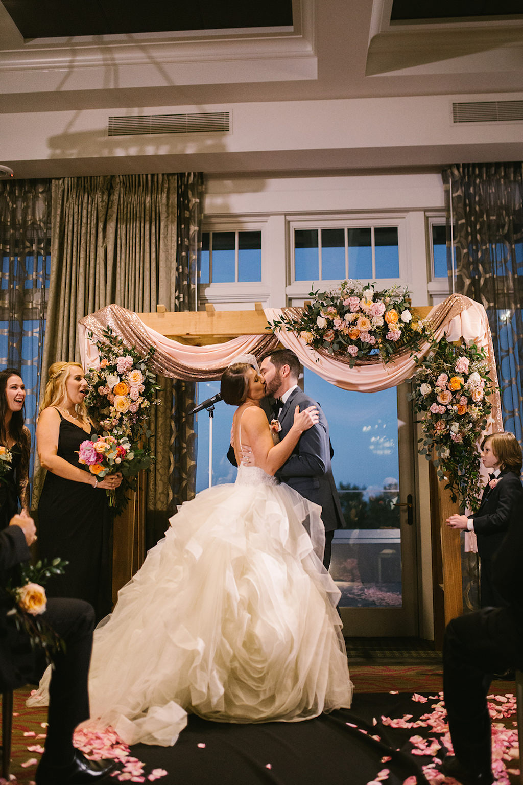 Tampa Bay Bride and Groom First Kiss Under Wooden Arch with Blush Pink and Sparkle Draping and Colorful Flower Bouquets Indoor Ballroom Wedding Ceremony Portrait | Downtown St. Pete Hotel Wedding Venue The Birchwood