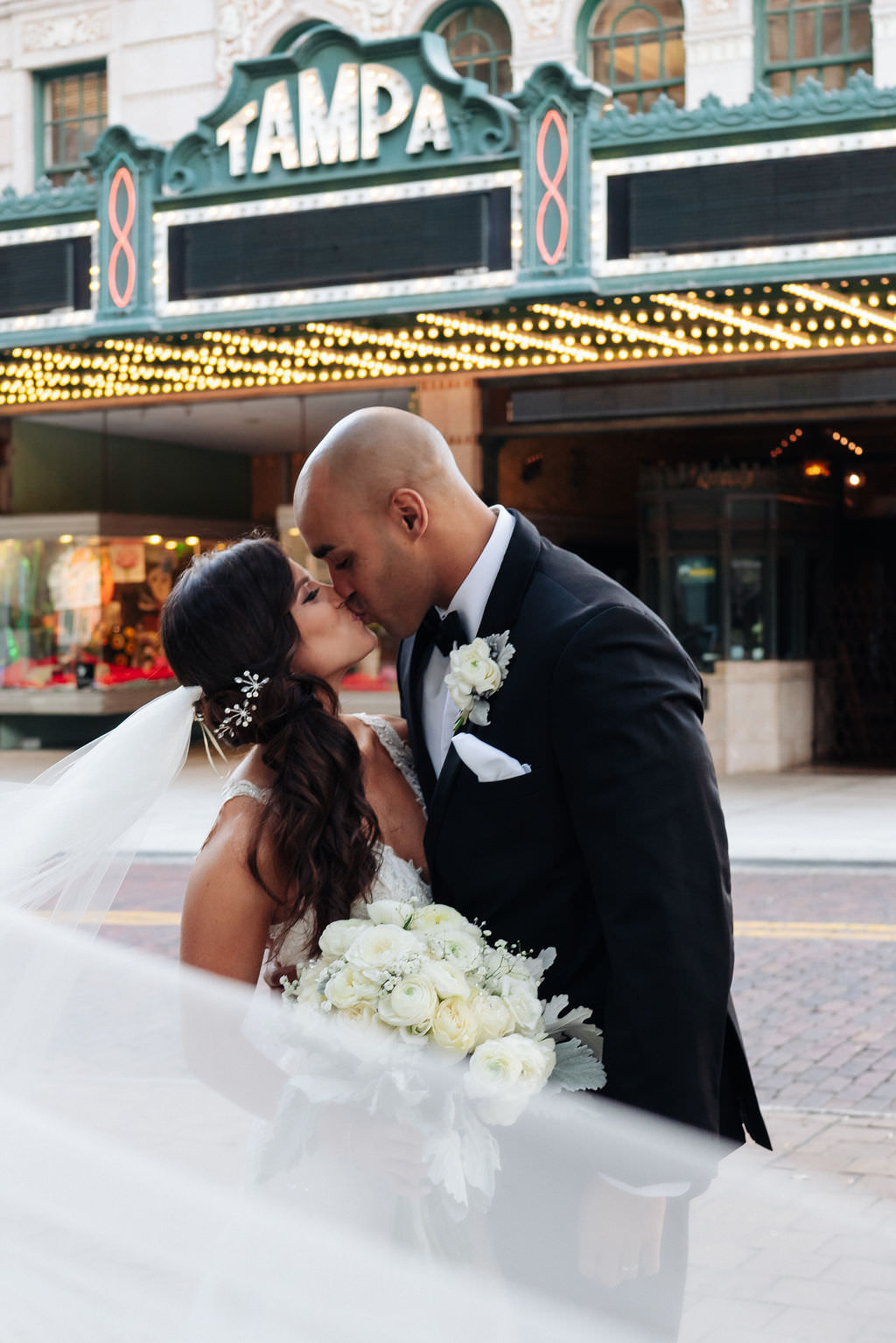 Florida Bride and Groom Intimate Wedding Portrait with Veil Flare, Kissing Outside of Historic Tampa Theatre, Carrying White and Ivory Rose Bouquet with Baby's Breath and Greenery | Photographer Grind & Press Photography