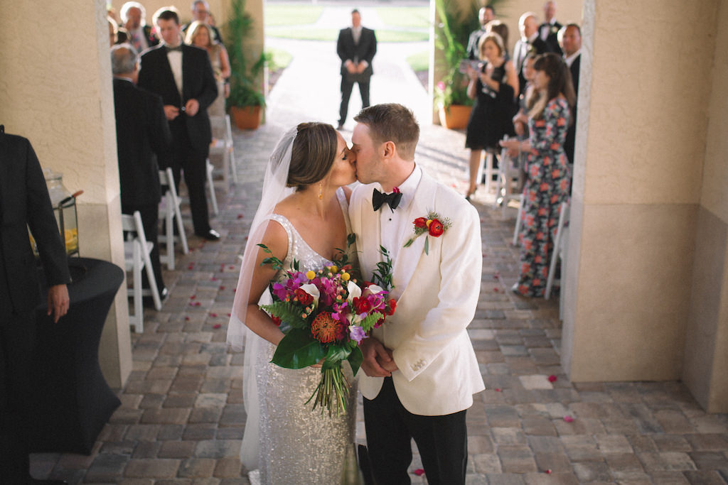 Bride and Groom First Kiss During Florida Wedding Ceremony, Bride in Sleeveless Dress Florals and Greenery, Groom in White Velvet Tuxedo Jacket with Red and Yellow Boutonniere | Tampa Bay Boutique Hotel Wedding Venue Fenway Hotel