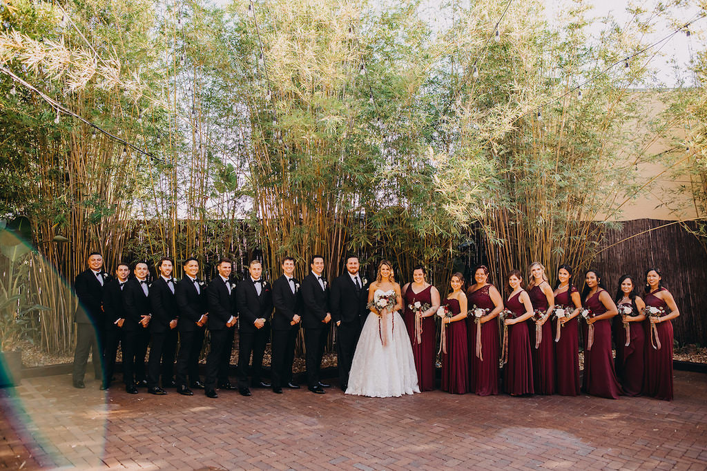 Florida Bride and Groom with Wedding Party in Romantic Outdoor Wedding Portrait in Romantic Bamboo Courtyard, Bride in a David's Bridal White Strapless Sweetheart Neckline Ball Gown Style Wedding Dress with Lace Overlay, Carrying Romantic Red Blush Pink and White Rose Wedding Bouquet with Greenery, Groom Wearing Classic Black Tux and Tie, Bridesmaids in Burgundy Wine Maroon Colored Mix and Match Style Long Dresses, Groomsmen in Black Tuxedo and Black Bowtie with Red Pocket Square | Downtown St. Pete Wedding Venue NOVA 535