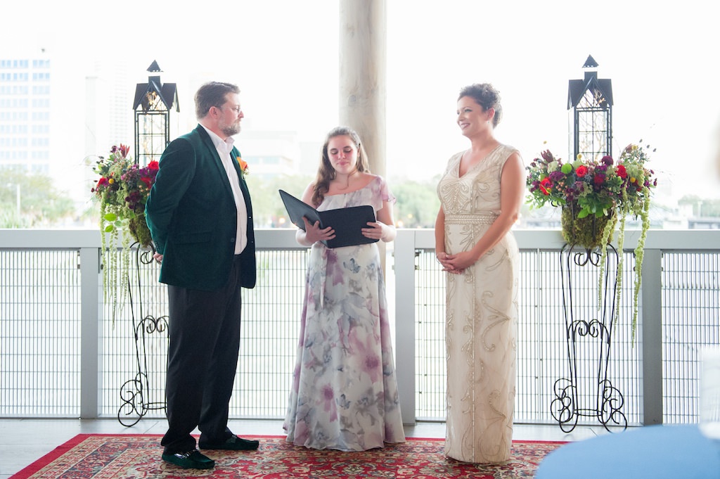 Tampa Bay Bride and Groom During Outside Waterfront Wedding Ceremony at Downtown Wedding Venue Tampa River Center with Child Officiant, Bride in Elegant Ivory and Gold Beaded Detailing V-Neck Sleeveless Sheath Style Wedding Dress, Groom in Elegant Jewel Tone Emerald Velvet Jacket, Romantic Ceremony and Alter Backdrop of Lantern in Garden Wedding Decor, Eclectic Colorful Mix Floral Wedding Flowers | Tampa Bay Wedding Photographer Andi Diamond Photography| Tampa Bay Wedding Makeup Artist Michelle Renee the Studio Tampa Bay Florist Apple Blossoms Floral Designs