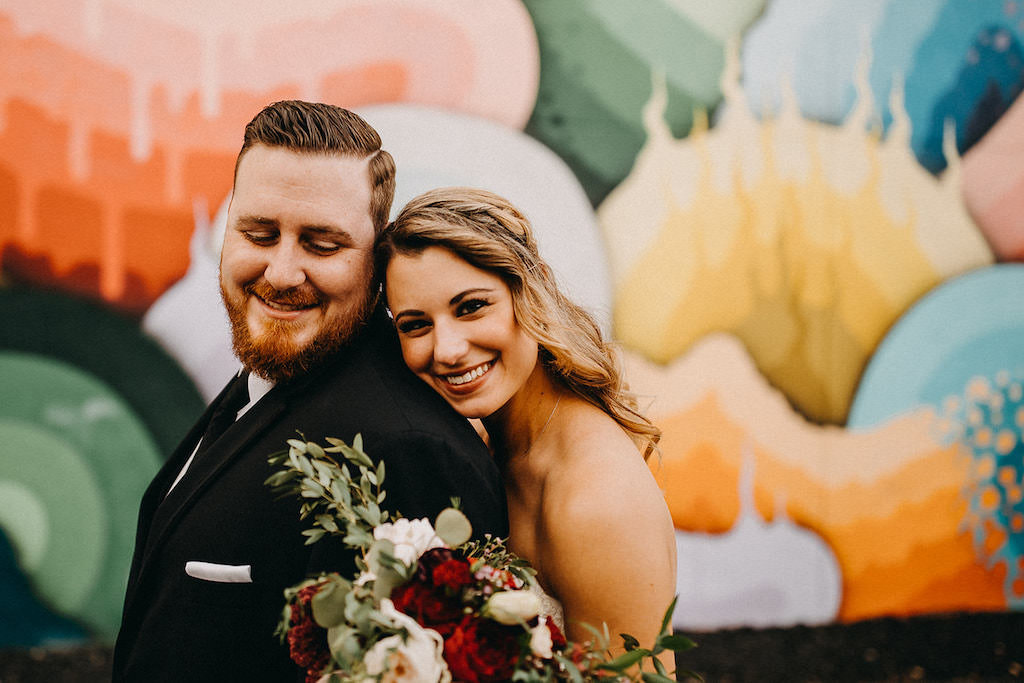 Bride and Groom Wedding Portrait in front of Colorful Art Mural, Bride With Half Up Half Down Braided Loose Curl Hairstyle, Carrying Romantic Red Blush Pink and White Rose Wedding Bouquet with Greenery at Tampa Bay Wedding, Groom in Classic Black Tuxedo with Black Tie | Downtown St. Pete