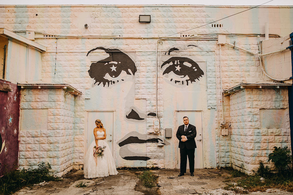 Tampa Bay Bride and Groom in Editorial Style Wedding Portrait in front of Artistic Art Wall, Bride Wearing David’s Bridal White Strapless Sweetheart Neckline Ballgown Style Wedding Dress, Carrying Romantic Red Blush Pink and White Wedding Bouquet with Greenery Wedding, Groom in Classic Black Tuxedo with Black Tie | Downtown St. Pete