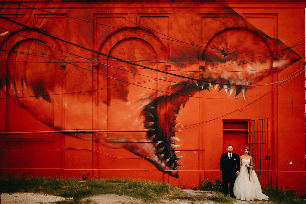 Tampa Bay Bride and Groom in Editorial Style Wedding Portrait in front of Artistic Red Shark Wall, Bride Wearing David’s Bridal White Strapless Sweetheart Neckline Ballgown Style Wedding Dress, Carrying Romantic Red Blush Pink and White Wedding Bouquet with Greenery Wedding, Groom in Classic Black Tuxedo with Black Tie | Downtown St. Pete