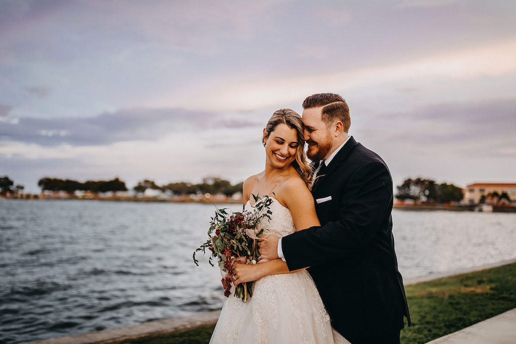 Tampa Bay Bride and Groom in Waterfront Wedding Portrait, Bride Wearing David’s Bridal White Strapless Sweetheart Neckline Ballgown Style Wedding Dress, With Half Up Half Down Braided Loose Curl Hairstyle, Carrying Romantic Red Blush Pink and White Rose Wedding Bouquet with Greenery at Tampa Bay Wedding, Groom in Classic Black Tuxedo with Black Tie | Downtown St. Pete Straub Park