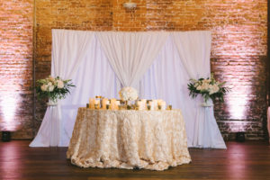 Classic Romantic Wedding Reception Decor, Sweetheart Table with Ivory Floral Linen, Candlesticks, Floral Bouquet Centerpiece, White Drapery and Brick Wall Backdrop, Greenery, Ivory, White Floral Bouquets on Clear Glass Pedestals | Photographer Kera Photography | Tampa Heights Industrial Wedding Venue Armature Works | Planner Kelly Kennedy Weddings and Events