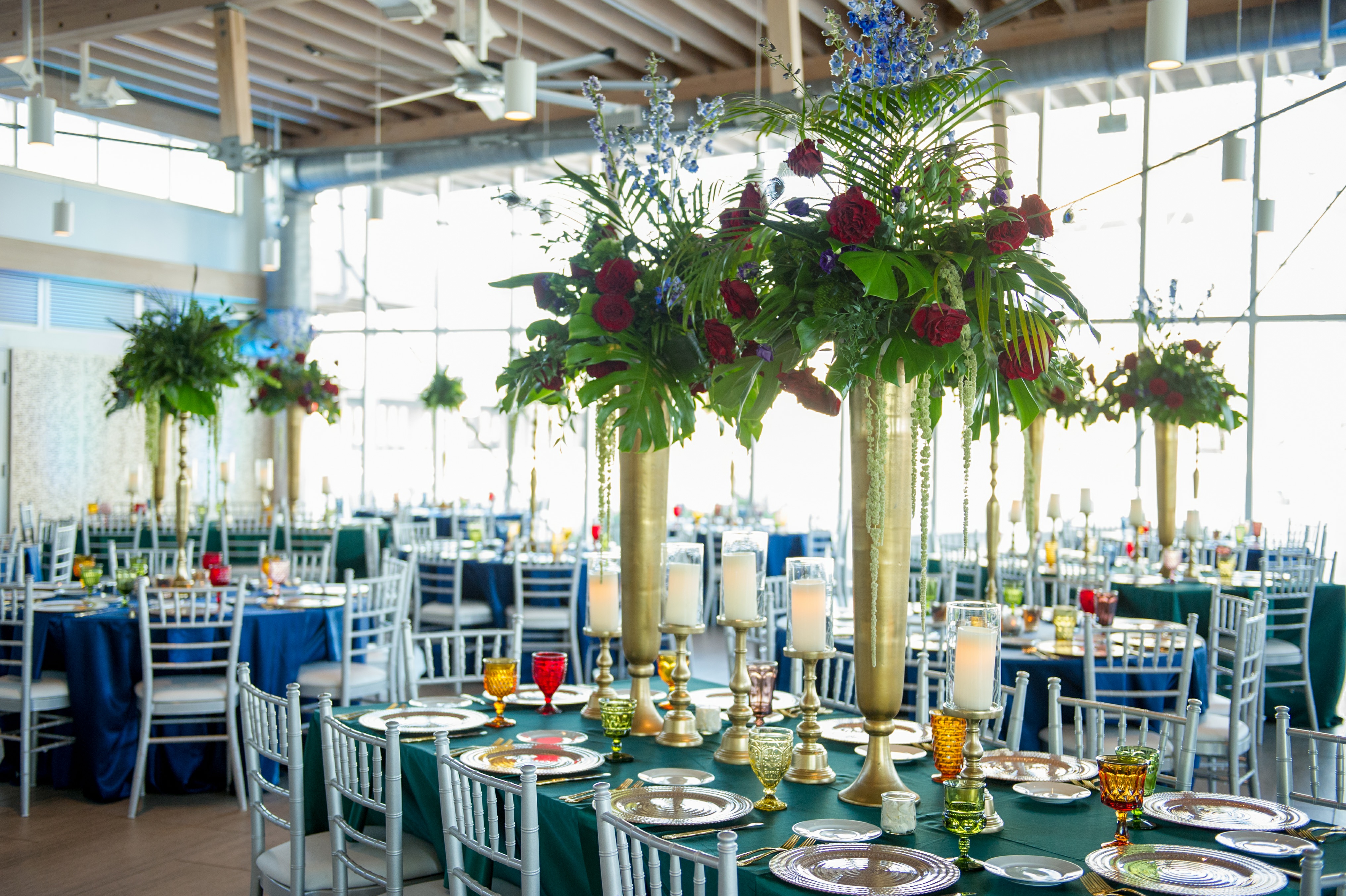 Elegant and Eclectic Jewel Toned Themed Reception Wedding Decor at Downtown Tampa Waterfront Reception Venue Tampa Riverwalk, Floor to Ceiling Windows, Large Gold Vase Centerpieces with Overflowing Green Floral and Red Rose Arrangements | Silk and Velvet Jewel Tone Tablecloths, Vintage Multi-colored Glass Goblets, Gold Flatware, Tall Silver Candelabras | Tampa Bay Wedding Photographer Andi Diamond Photography