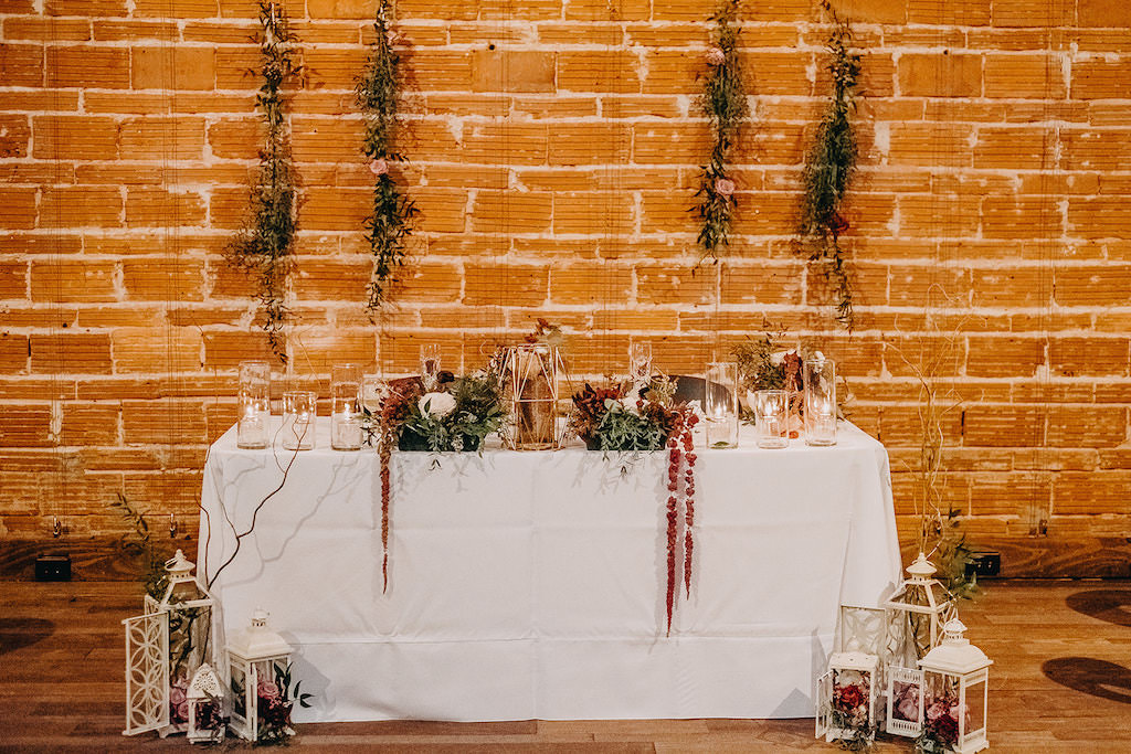 Romantic Boho White Sweetheart Table, Vintage White Lanterns with Red Roses and Glass Liquid Candles, Hanging Greenery and Roses in front of Red Exposed Brick Wall | Downtown St. Pete Wedding Venue NOVA 535