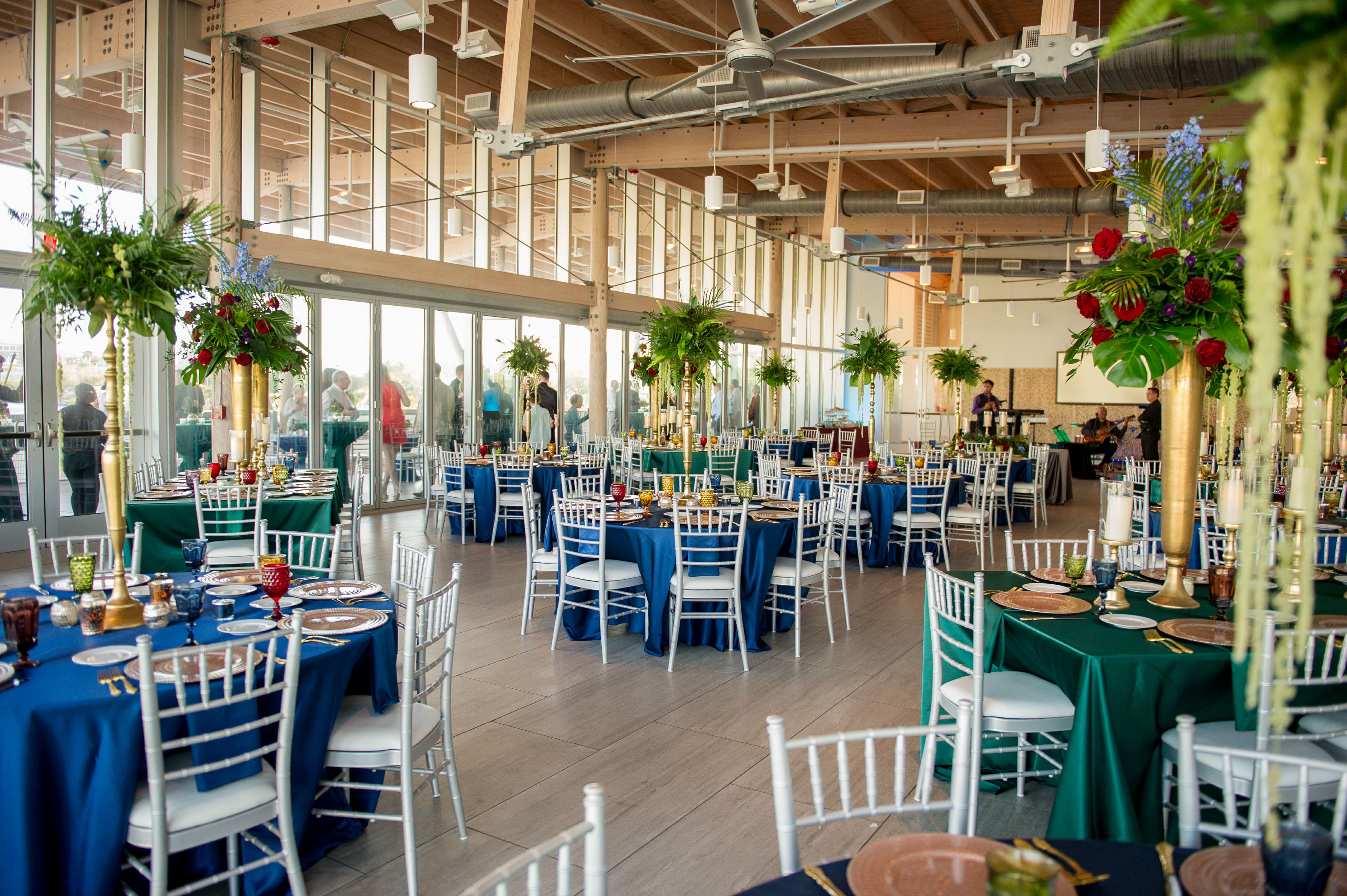 Elegant and Eclectic Jewel Toned Themed Reception Wedding Decor at Downtown Tampa Waterfront Reception Venue Tampa River Center, Floor to Ceiling Windows, Large Gold Vase Centerpieces with Overflowing Green FloralArrangements | Silk and Velvet Jewel Tone Tablecloths, Silver Chairs, Vintage Multi-colored Glass Goblets, Gold Flatware, Tall Silver Candelabras | Tampa Bay Wedding Photographer Andi Diamond Photography