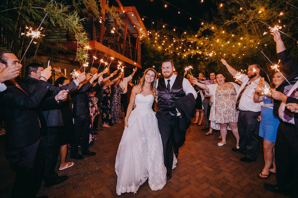 Tampa Bay Bride and Groom During Wedding Sparkler Exit in Outdoor Bamboo Courtyard with Romantic String Lights, Bride in David's Bridal White Strapless Sweetheart Neckline Ballgown Style Wedding Dress with Lace Overlay, Groom in Black Vest and Tie | St. Pete Wedding Venue NOVA 535