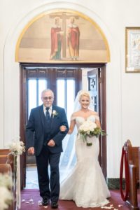 Florida Bride and Father of the Bride During Wedding Processional in Cathedral Ceremony, Bride Wearing Pronovias White Lace Wedding Dress with Sweetheart Neckline Fitted Bodice and Side Lace Illusion Sleeves, Carrying Elegant White Rose Bridal Bouquet with Greenery | Tampa Bay Bridal Dress Shop Nikki and Glitz Glam Boutique