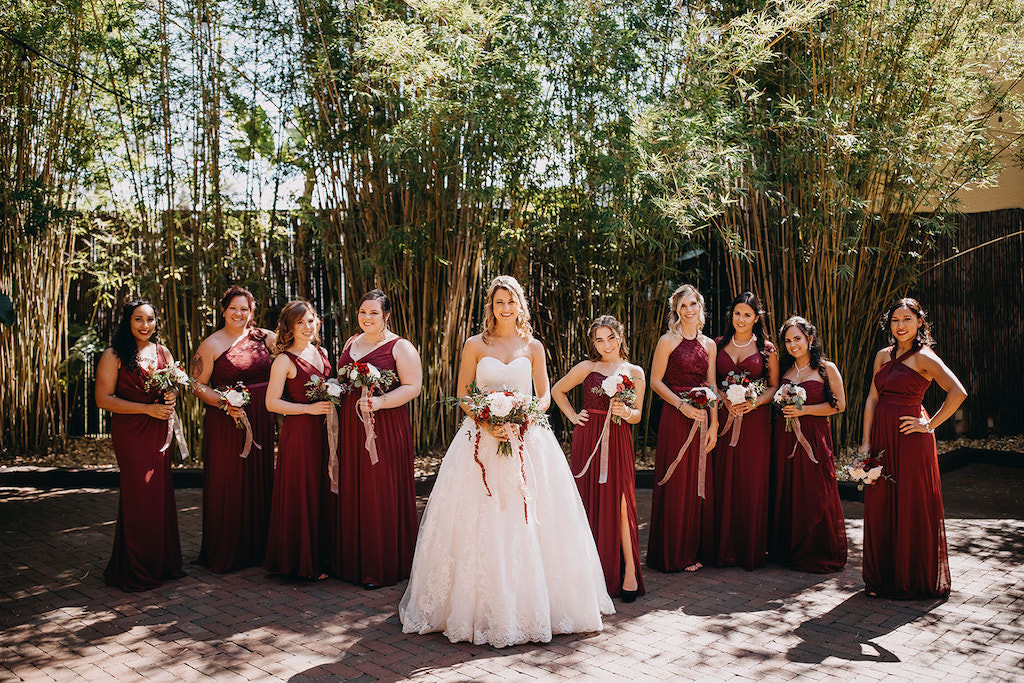 Florida Bride and Bridesmaids Bridal Party Portrait in Romantic Outdoor Bamboo Courtyard, Bride in a David's Bridal White Strapless Sweetheart Neckline Ball Gown Style Wedding Dress with Lace Overlay, Carrying Romantic Red Blush Pink and White Rose Wedding Bouquet with Greenery, Bridesmaids in Burgundy Wine Maroon Colored Mix and Match Style Long Dresses | Downtown St. Pete Wedding Venue NOVA 535